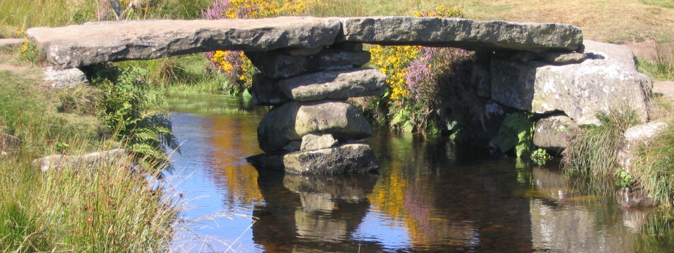 Ancient stone clapper bridge over a calm stream with flowering gorse and heather, surrounded by grass in earthy green and yellow tones